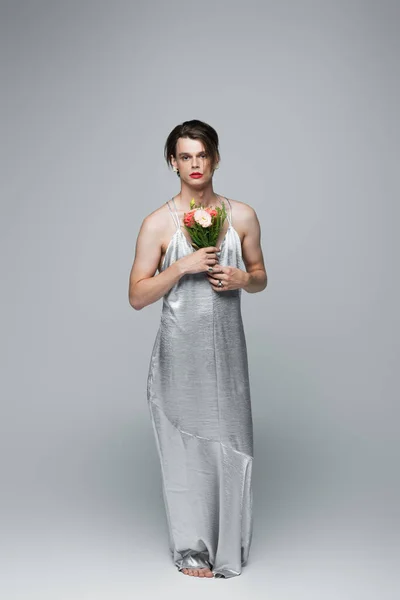 full length of transgender man in slip dress posing with flowers on gray
