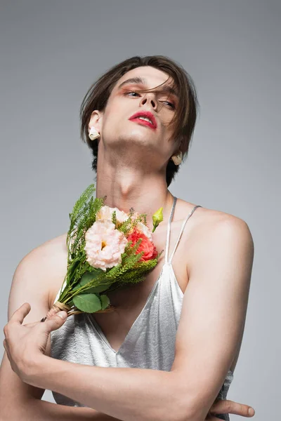 young transgender man in slip dress posing with flowers isolated on gray