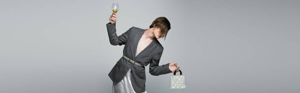 трансгендерный мужчина в платье с блейзером держа кошелек и бокал вина изолированы на сером, баннер