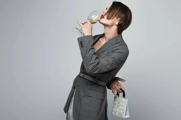 transgender man in blazer drinking wine while holding purse isolated on gray