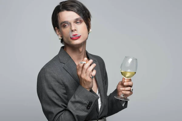 young transgender man in blazer and earrings holding glass of wine while smoking isolated on grey