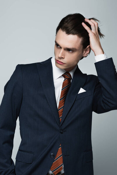 young androgynous man in blazer and tie adjusting hair isolated on grey