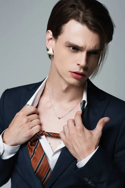 スーツの若いトランスジェンダーのビジネスマンがシャツを調整しグレーで隔離されたネックレスを見せる — ストック写真