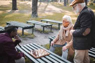 Parktaki satranç tahtasının yanındaki Afrikalı Amerikalı arkadaşa bakan ırklar arası adamlar. 