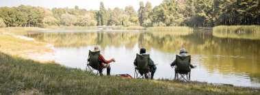 Multiethnic elderly friends in fishing outfit sitting on chairs near lake, banner  clipart