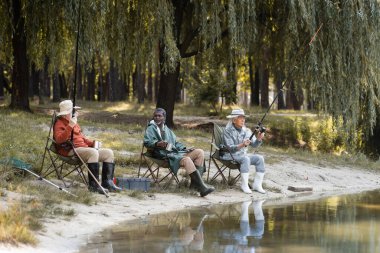 Çok ırklı yaşlı adamlar ellerinde olta çubuklarıyla göl kenarında termo bardaklar tutuyorlar. 