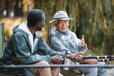 Kıdemli Asyalı adam dışarıda balık tutarken Afrikalı Amerikalı arkadaşıyla konuşuyor. 