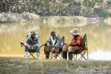 Balıkçı kıyafetli ırklar arası pozitif adamlar göl kıyısındaki sandalyelerde termo bardakları tutuyorlar. 