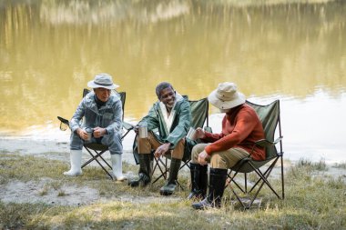 Şapkalı ve balıkçı kıyafetli ırklar arası kıdemliler göl kenarındaki sandalyelerde termo bardakları tutuyorlar. 