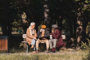 Arkadaşlarının yanında oturan kıdemli ırklar arası adamlar parkta akıllı telefondan konuşuyorlar. 