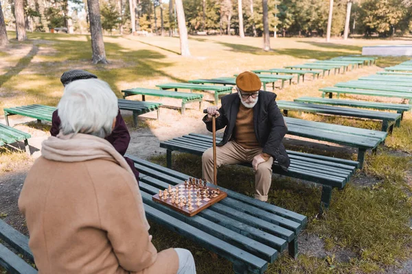 Man with walking cane sitting near friends and chess in park