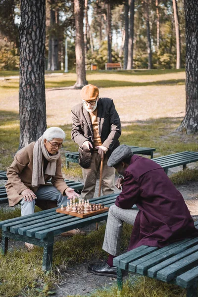 公園でチェスをしている多文化の友人の近くに立って杖を歩くシニア男 — ストック写真