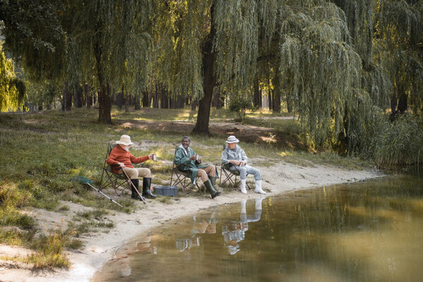 Smiling interracial senior men in rubber boots and fishing outfit holding cups near lake in park 