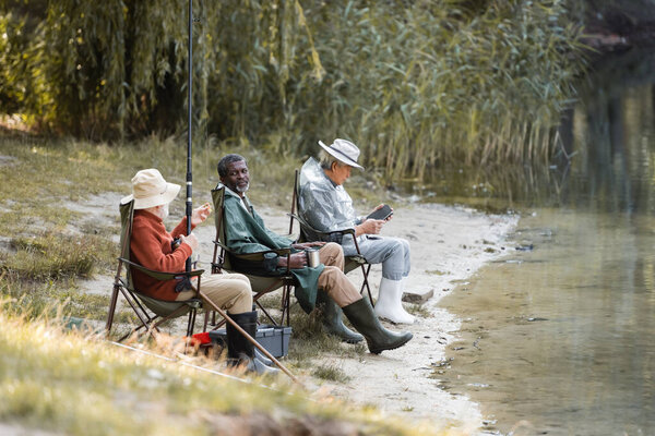 African american man holding thermo cup near interracial friends fishing near lake 