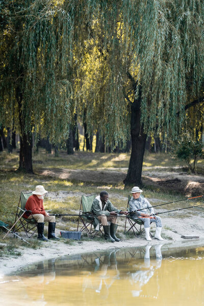 Cheerful multicultural men in rubber boots fishing on chairs near lake in park 