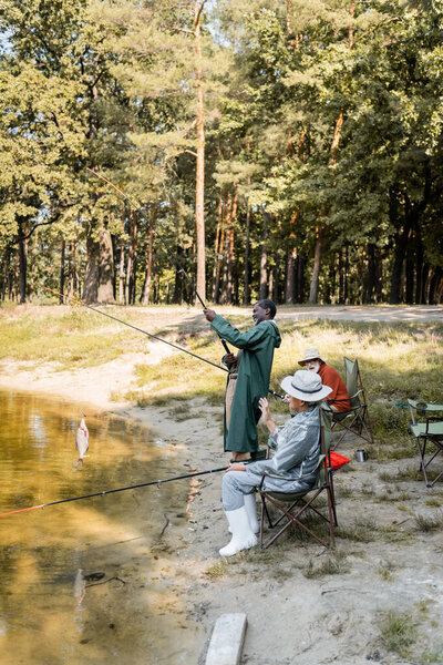 Multicultural senior men fishing in lake near excited friend in park 