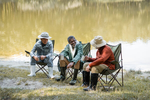 Interracial senior men in hats and fishing outfit holding thermo cups on chairs near lake 