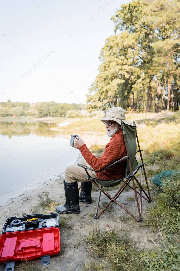 Senior man in fishing outfit holding thermo cup near toolbox and lake 
