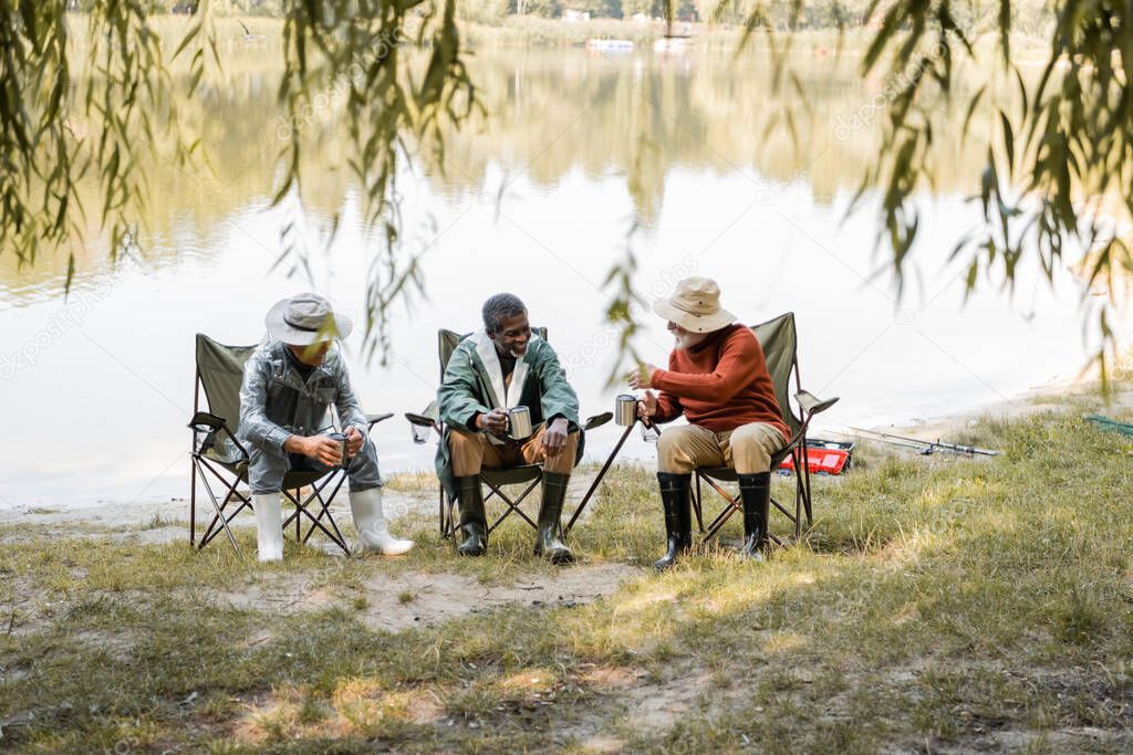 Senior man with thermo cup talking to interracial friends in fishing outfit on lake coast 