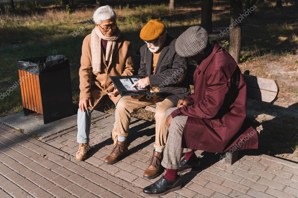 Senior man pointing at photo in album near interracial friends in autumn park