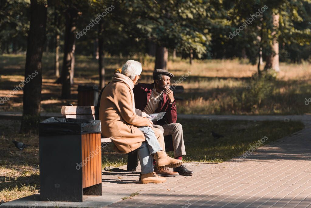African american man sitting near friends with photo album in park during autumn