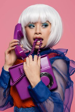 Asian pop art model biting cable while talking on telephone isolated on pink  clipart