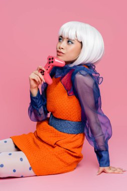 KYIV, UKRAINE - 10 Aralık 2020: Pembe arka planda otururken güzel Asyalı pop sanat modeli joystick 'i tutuyor