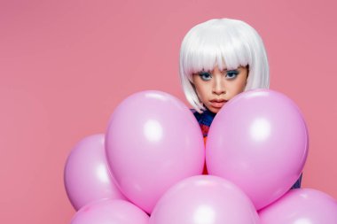 Beyaz peruklu genç Asyalı kadın pembe balonların yanındaki kameraya bakıyor. 