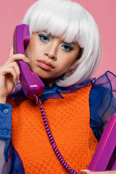 Young asian pop art woman looking at camera while talking on telephone isolated on pink 
