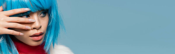 Young asian woman in wig looking away and touching face isolated on blue, banner 