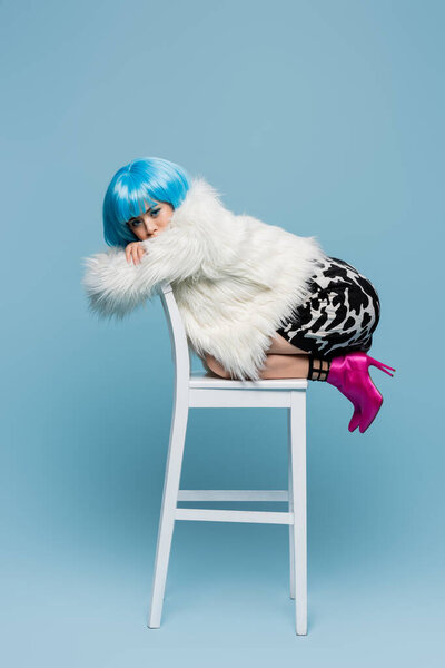 Stylish asian woman in heeled shoes and fluffy jacket posing on chair on blue background 