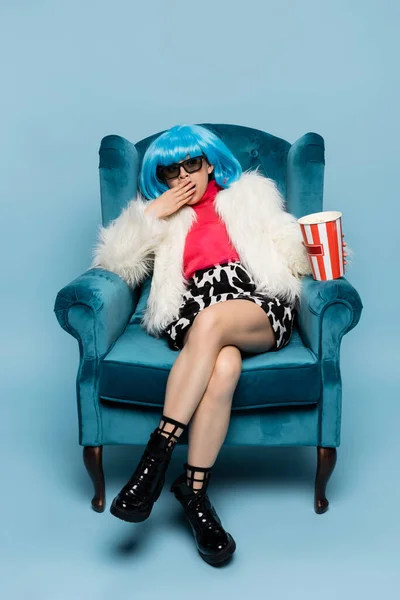 Stylish asian pop art woman yawning and holding popcorn on armchair on blue background