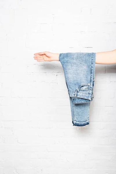 Jeans azul pendurado na mão feminina perto da parede de tijolo branco no fundo — Fotografia de Stock