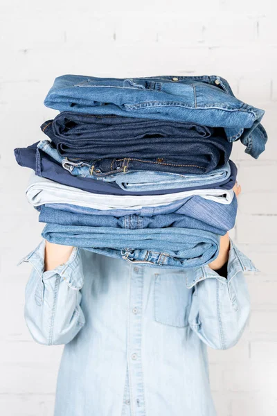 Partial view of woman obscuring face with stack of denim clothes near white brick wall — Stock Photo