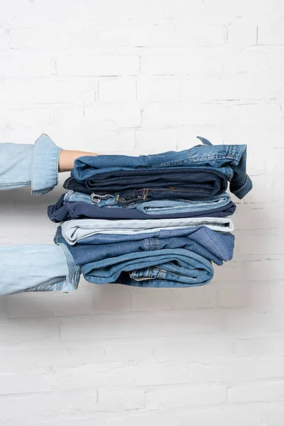 Partial view of woman holding stack of blue denim clothes near white brick wall — Stock Photo