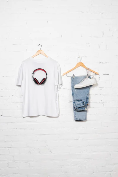 T-shirt, fones de ouvido sem fio, jeans azuis e sapatilha em cabides perto da parede de tijolo branco — Fotografia de Stock