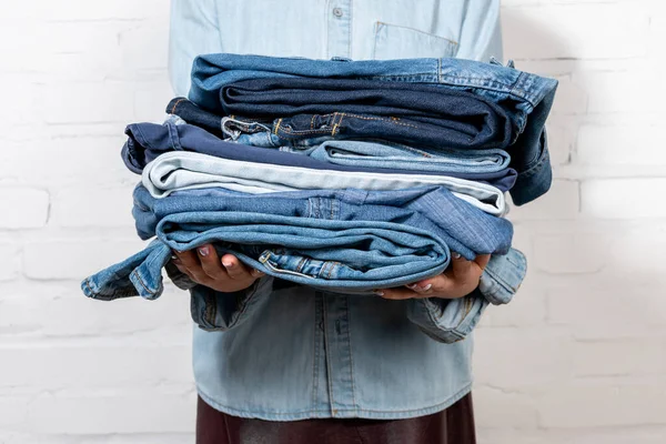 Vista recortada de la mujer sosteniendo pila de ropa de mezclilla azul cerca de la pared de ladrillo blanco - foto de stock