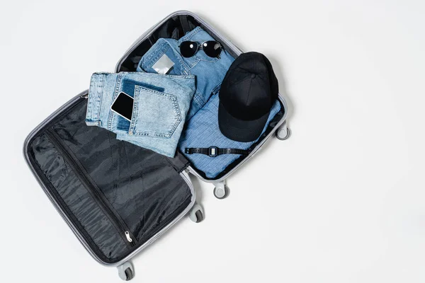 Vue du dessus de la valise remplie de vêtements en denim, casquette noire, smartphone, lunettes de soleil et montre-bracelet électronique sur blanc — Photo de stock