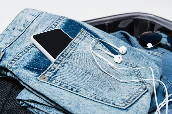 Закрытый вид смартфона в кармане джинсов рядом с наушниками и солнцезащитными очками в дорожной сумке — стоковое фото