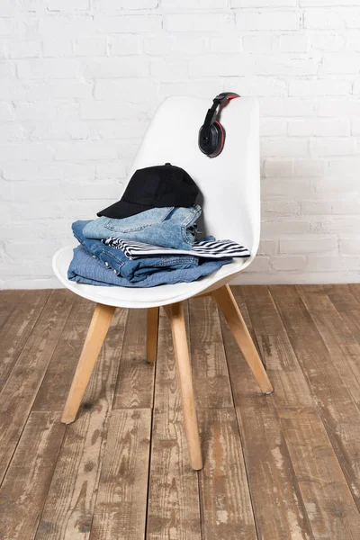 Silla con pila de jeans, gorra negra y auriculares inalámbricos en suelo de madera cerca de la pared de ladrillo blanco - foto de stock