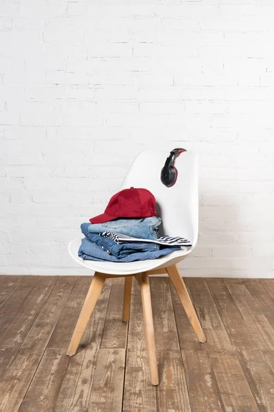 White chair with stacked denim clothes, burgundy cap and wireless headphones on wooden floor near brick wall — Stock Photo