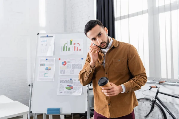 Hombre de negocios árabe hablando por celular y sosteniendo café para acercarse al rotafolio en la oficina - foto de stock