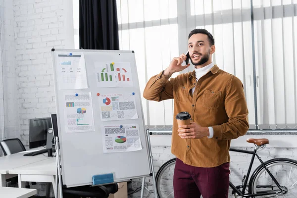 Smiling muslim businessman talking on smartphone and holding takeaway drink in office — Stock Photo