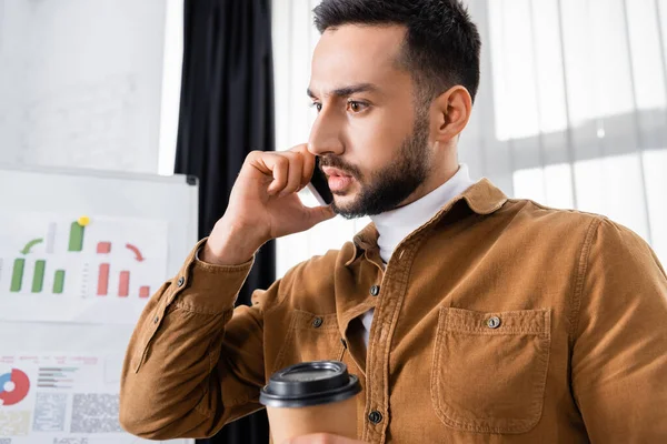 Homme d'affaires musulman parlant sur smartphone et tenant du café pour aller au premier plan flou — Photo de stock