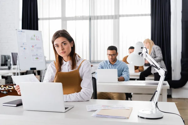 Imprenditrice che lavora con documenti e laptop vicino a colleghi multietnici su sfondo sfocato — Foto stock