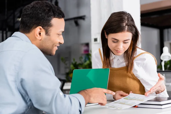 Mujer de negocios señalando el documento cerca de un colega indio sonriente en un primer plano borroso - foto de stock