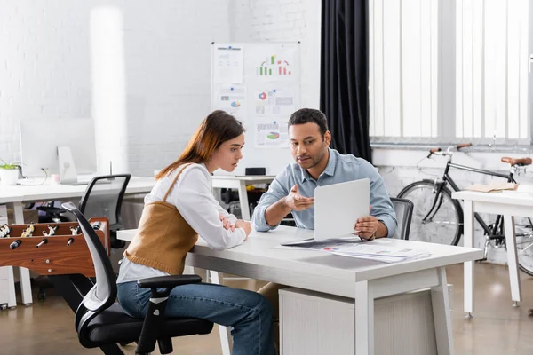 Hombre de negocios indio señalando portátil cerca de colega en la oficina - foto de stock