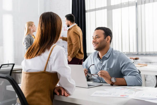 Hombre de negocios indio sonriente mirando a su colega cerca de papeles y portátil en primer plano borroso - foto de stock