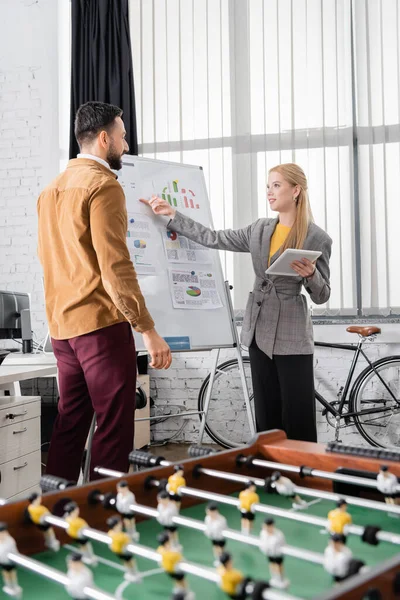 Smiling businesswoman with digital tablet pointing on flipchart near arabian colleague and table soccer on blurred foreground — Stock Photo