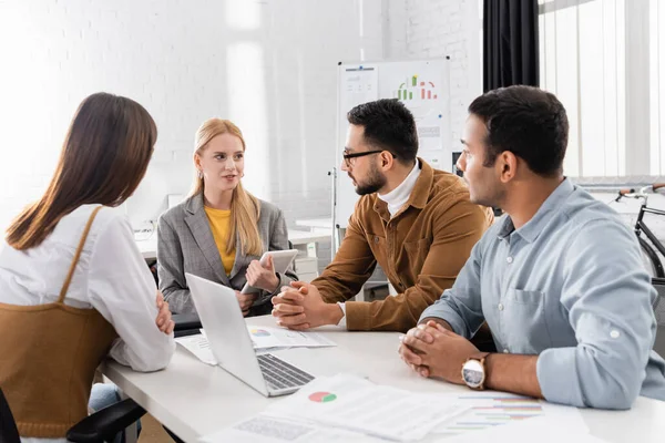 Smiling businesswoman with digital tablet looking at multiethnic colleagues near papers on blurred foreground — Stock Photo